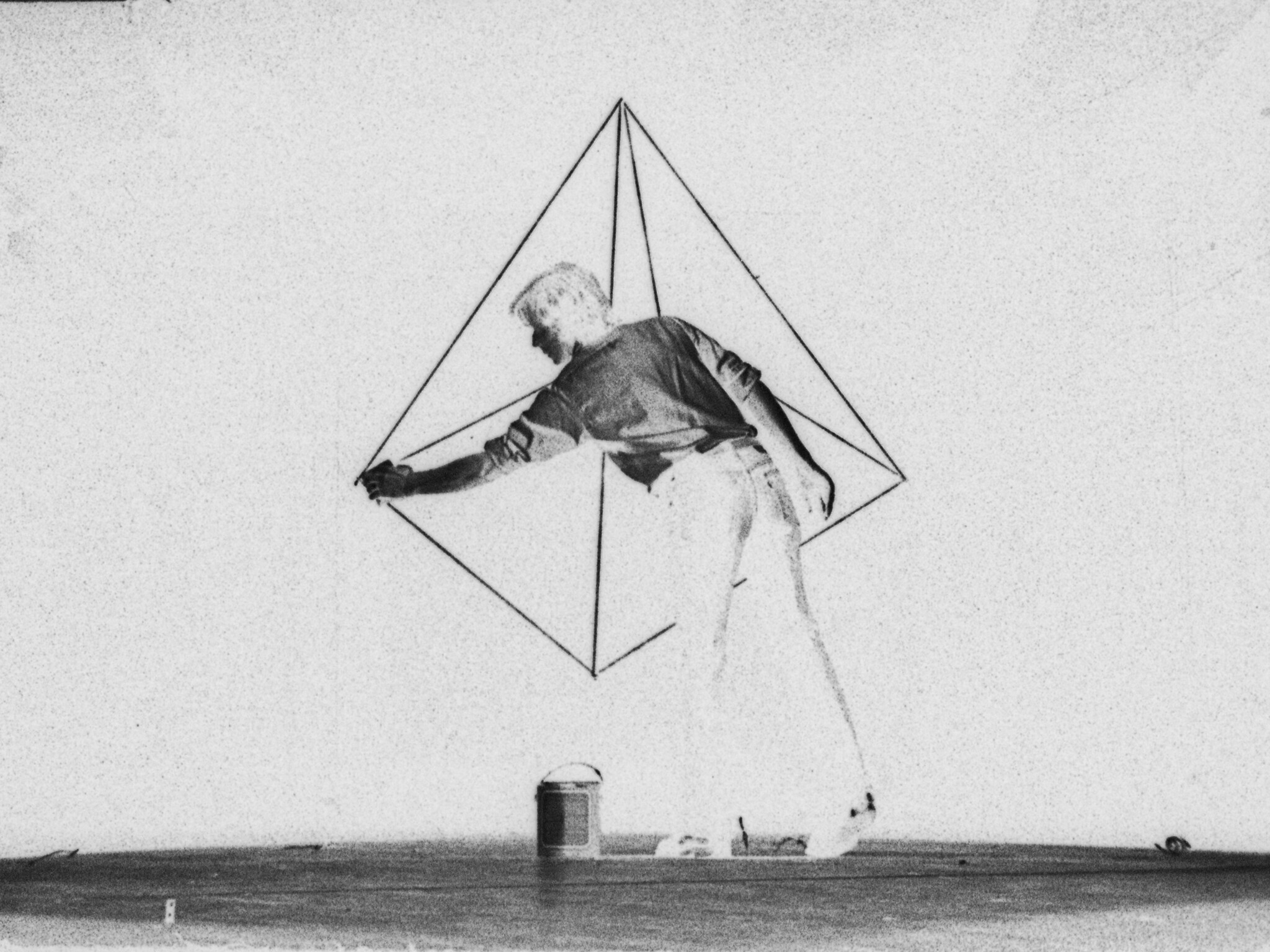 A person stands in front of a large geometric wire shape, reaching out with one arm to touch it.
