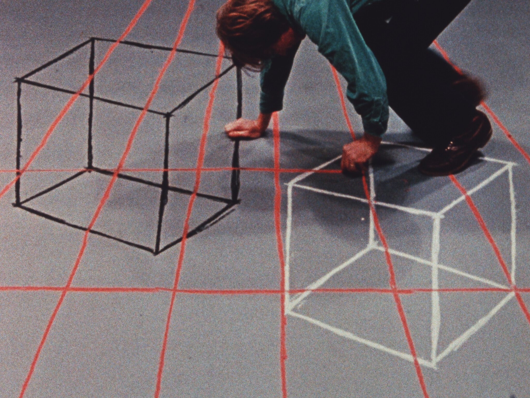 Person crouching on a floor with red grid lines, drawing two cubes with black and white outlines.