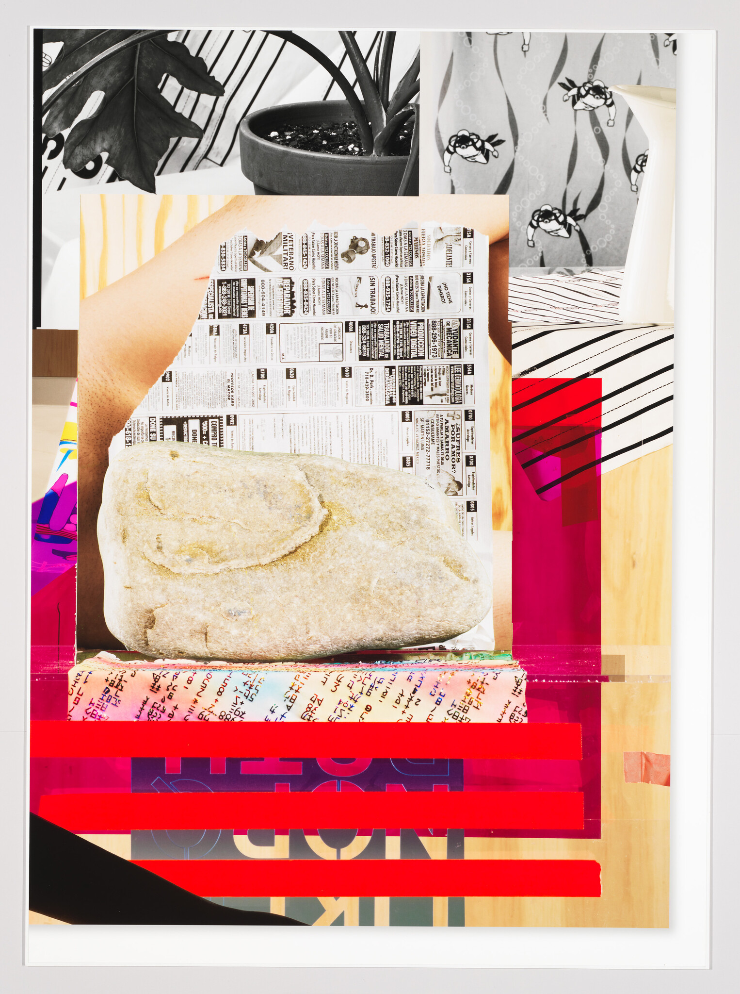 A large stone rests on folded newspapers and colorful layered papers with red tape.