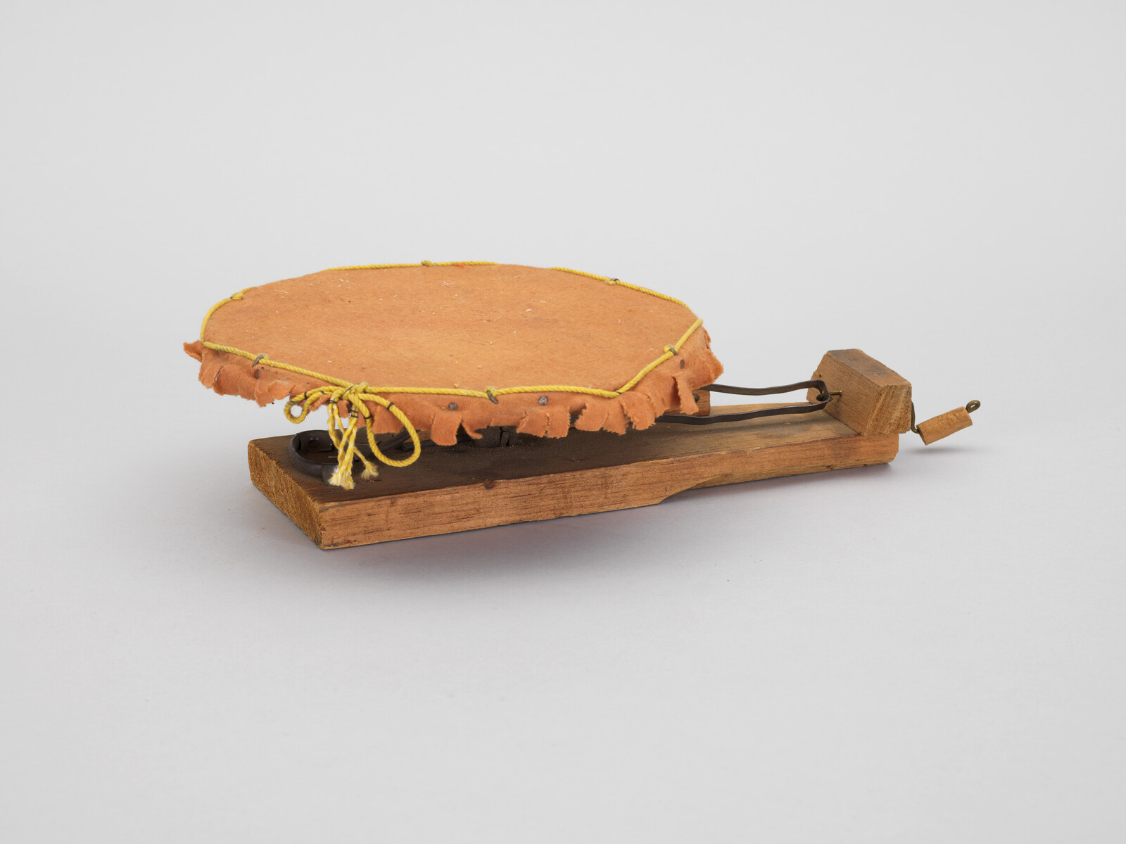 Small handheld drum with stretched leather head tied by yellow cord mounted on wooden base.