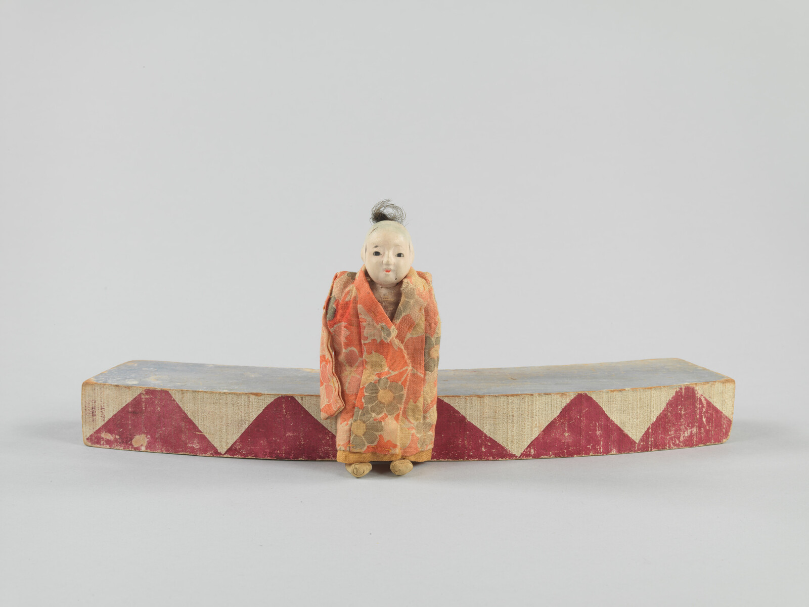 Small traditional Japanese doll in a patterned kimono stands in front of a curved wooden block with red triangles.