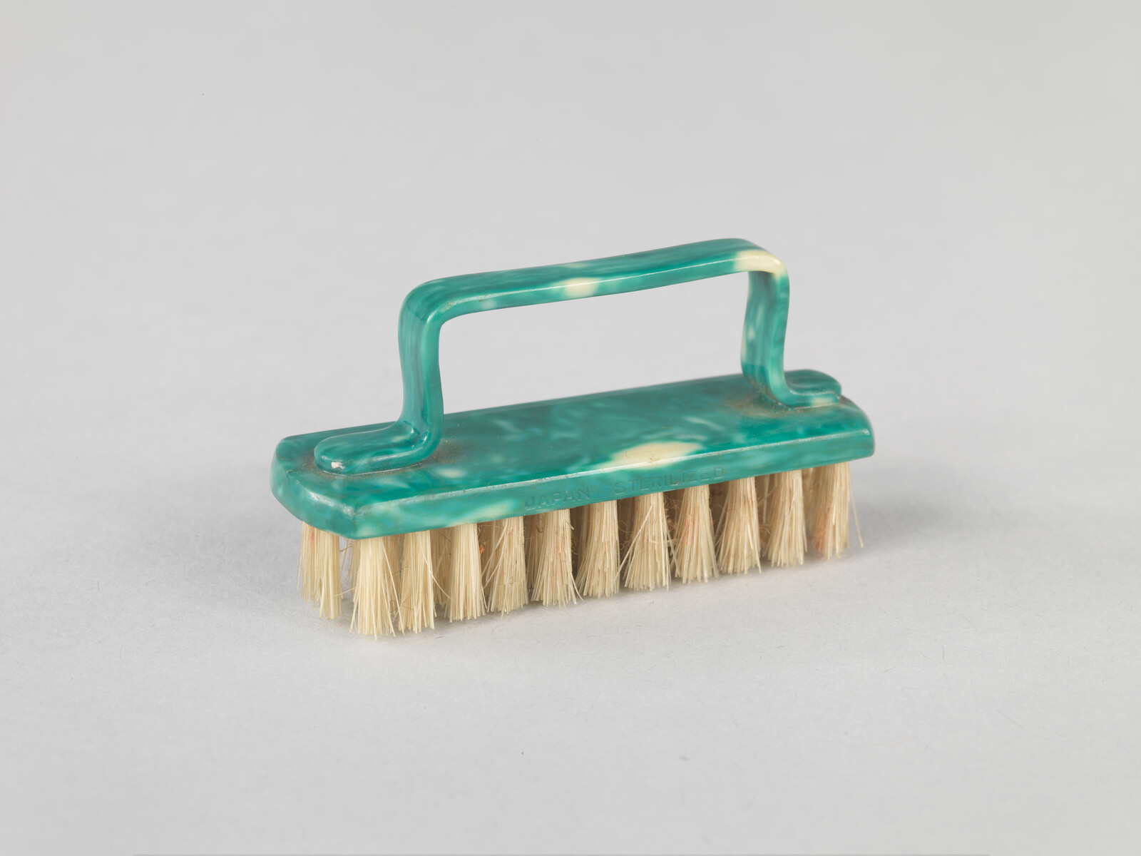 Green handled scrub brush with beige bristles, resting on a light gray surface.