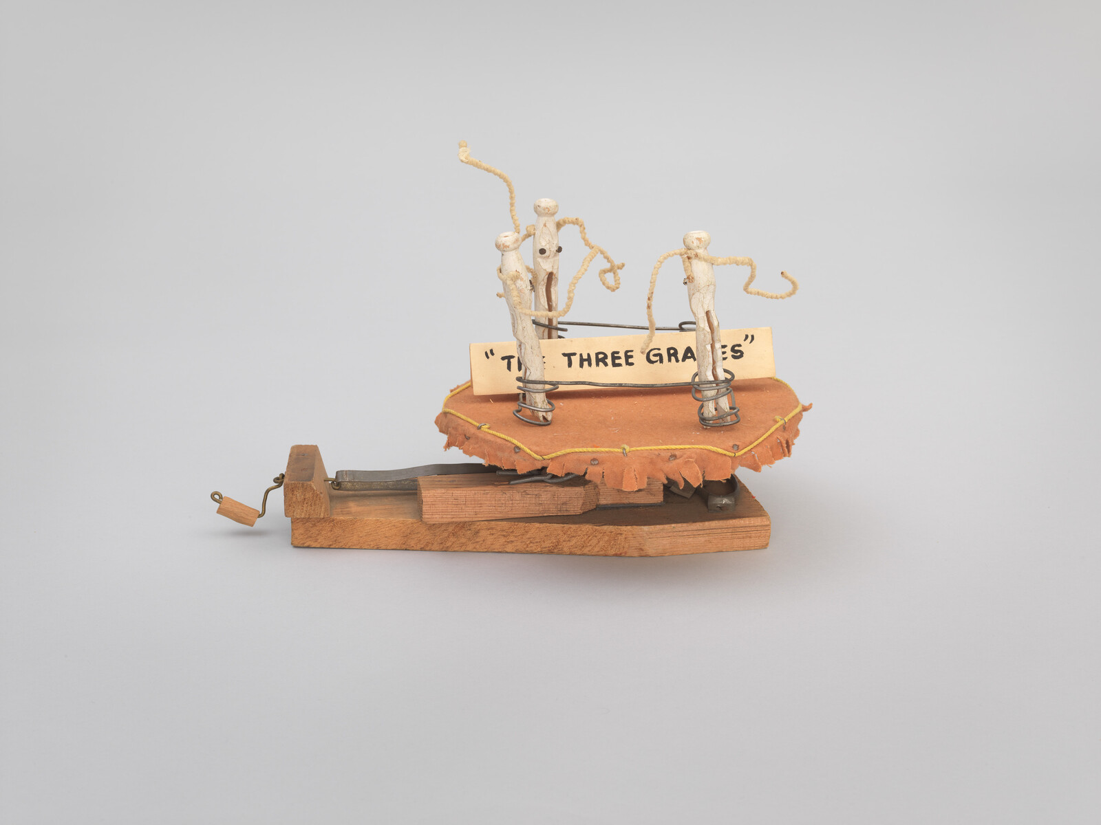 A small wooden automaton titled "The Three Graces" with three wire-and-wood figures on a leather platform.