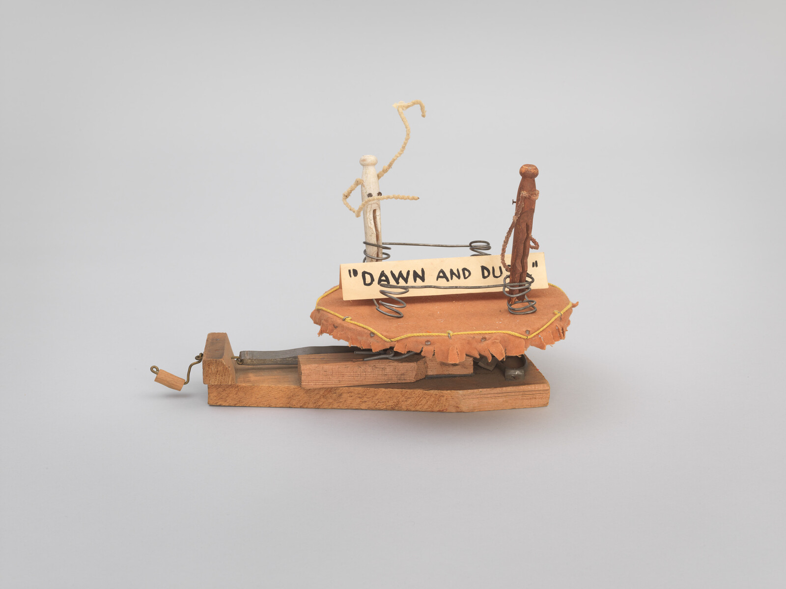 Two simple wire and wood figures stand on a small stage labeled "DAWN AND DUSK" with a hand crank.