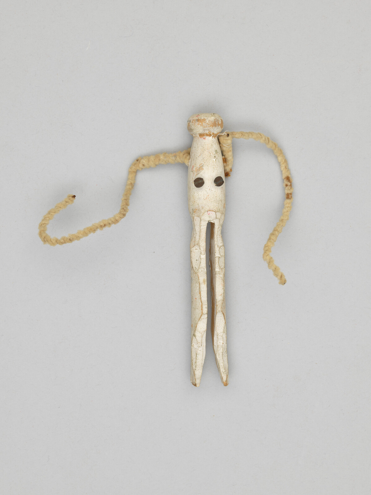 Carved white wooden figure with two-pronged legs, dark round eyes, and braided rope arms.