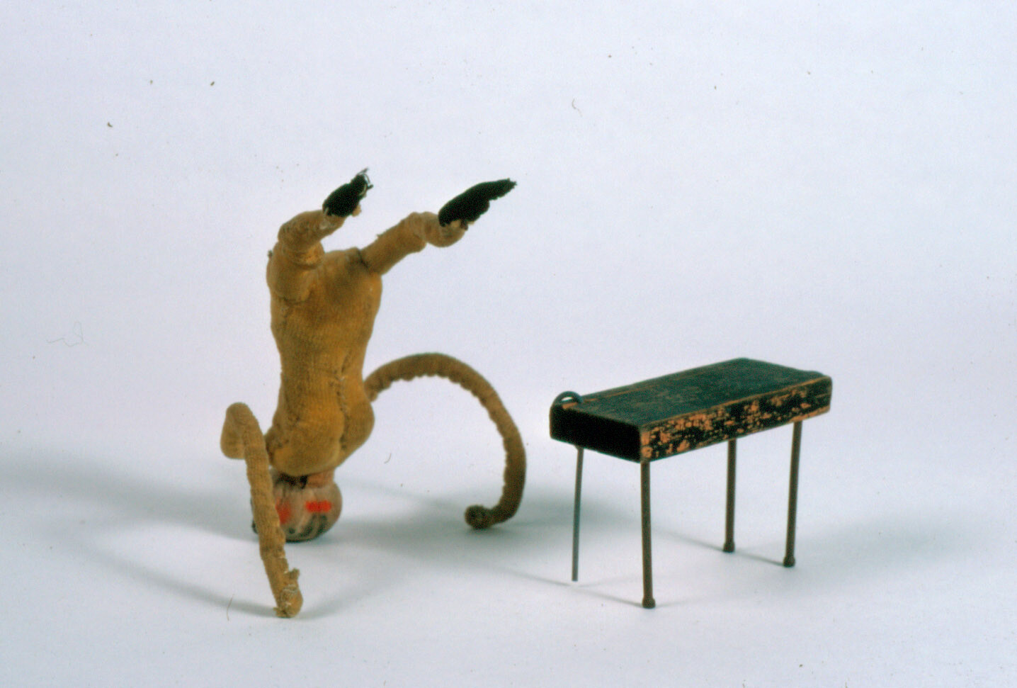 Small stuffed monkey doing a headstand next to a worn miniature gym pommel horse.