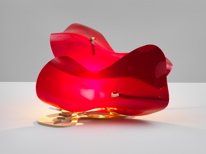 A glossy red abstract sculpture with flowing curves mounted on a wooden base under soft light.