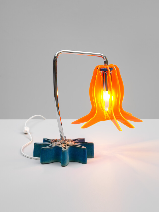 A modern table lamp with orange petal-like shade, chrome arm, and blue star-shaped base with pebbles.