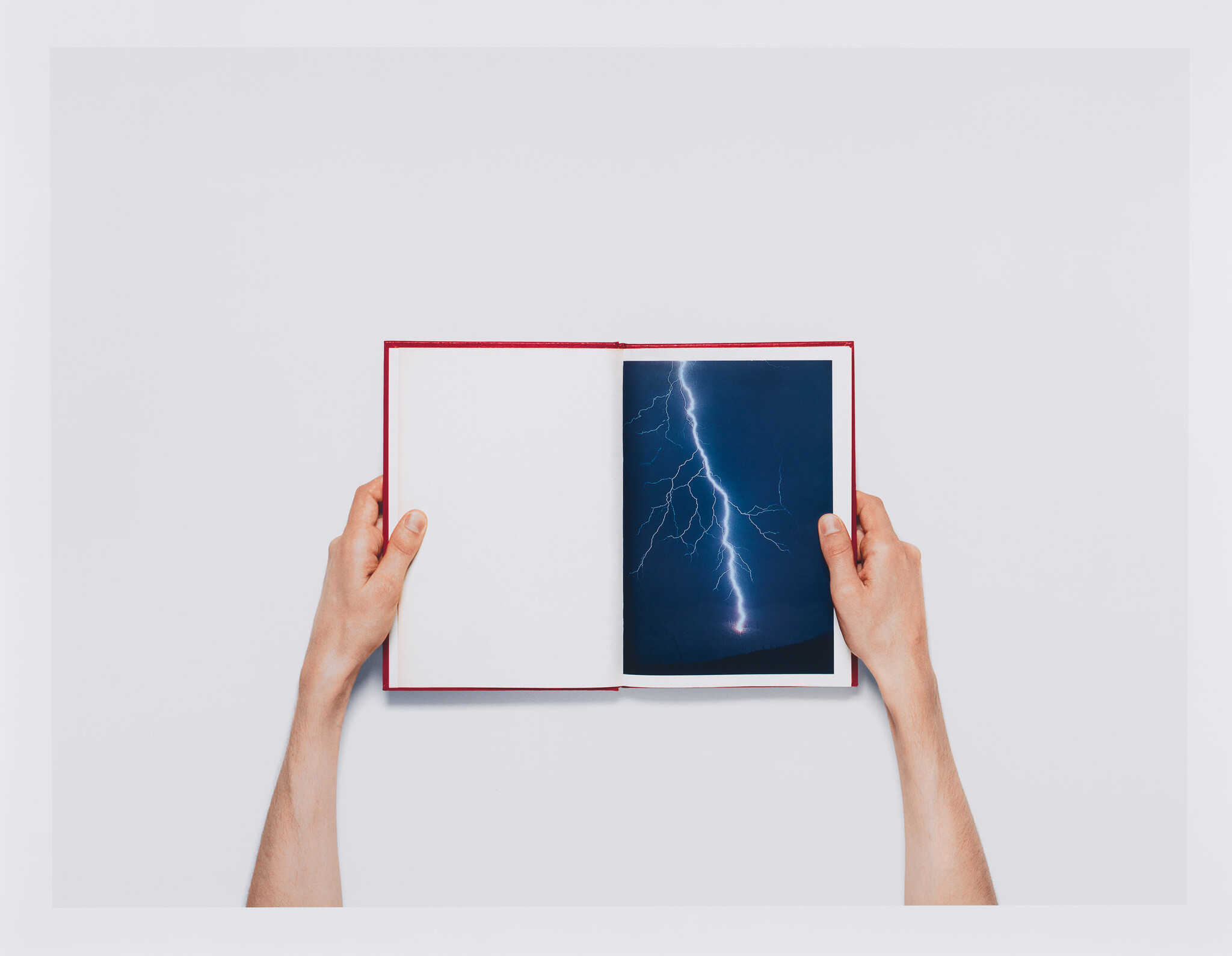 Hands hold an open book showing a blank page and a photograph of lightning.