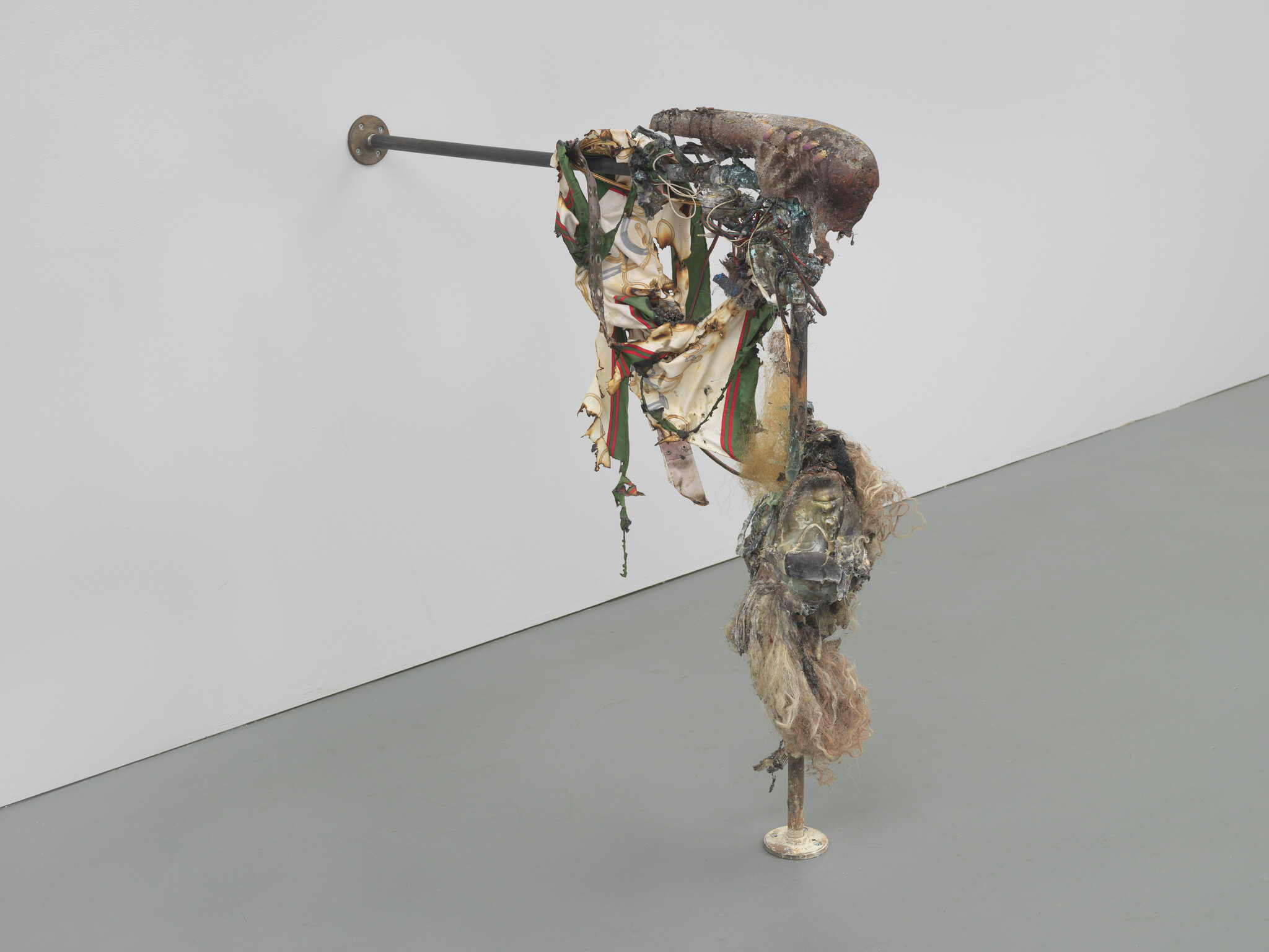 Sculpture of rusted metal and torn fabric hanging from a wall rod with a distorted face.