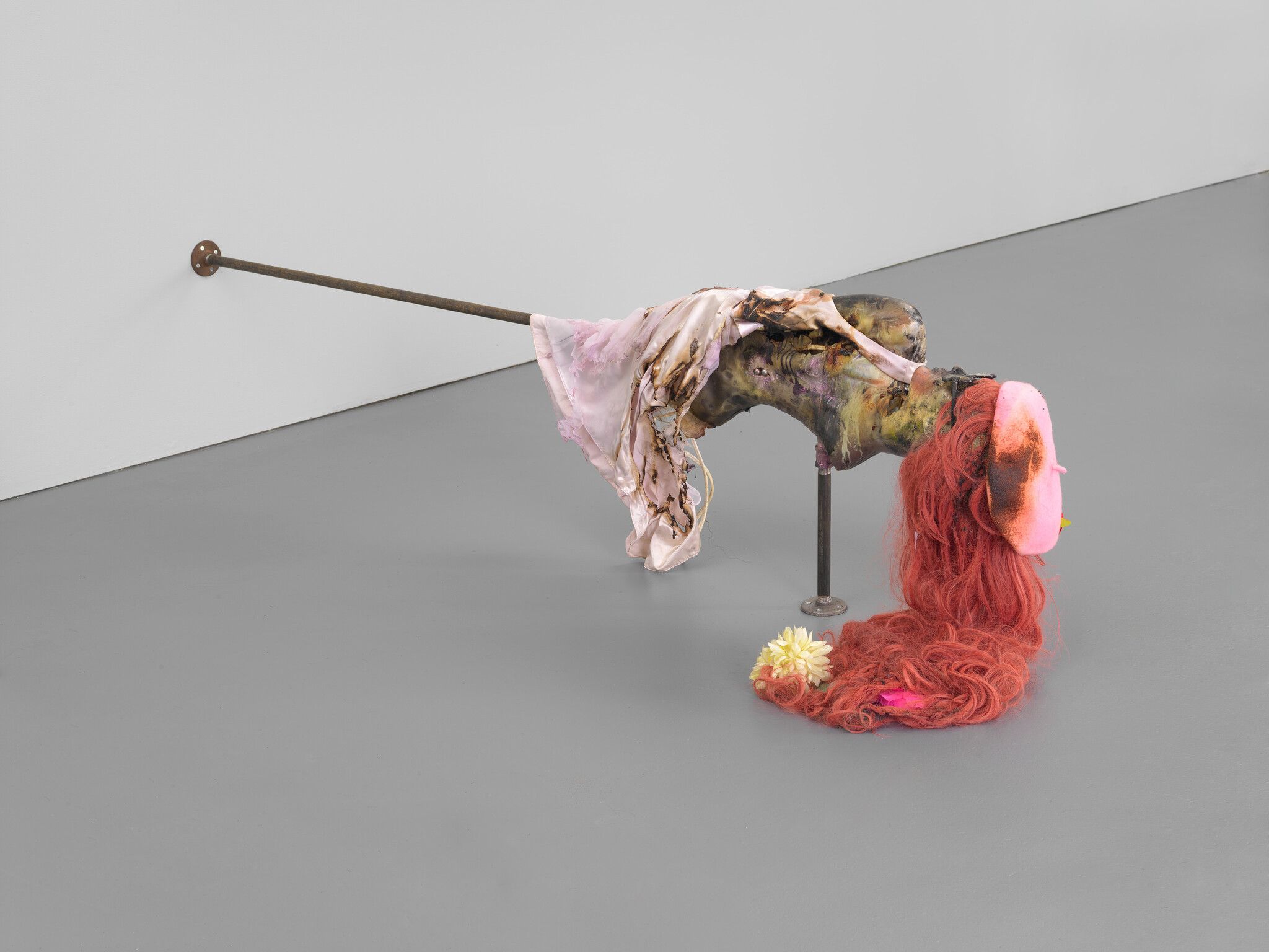 A sculpted torso with burned fabric and long red hair hangs from a metal pole in a gallery.