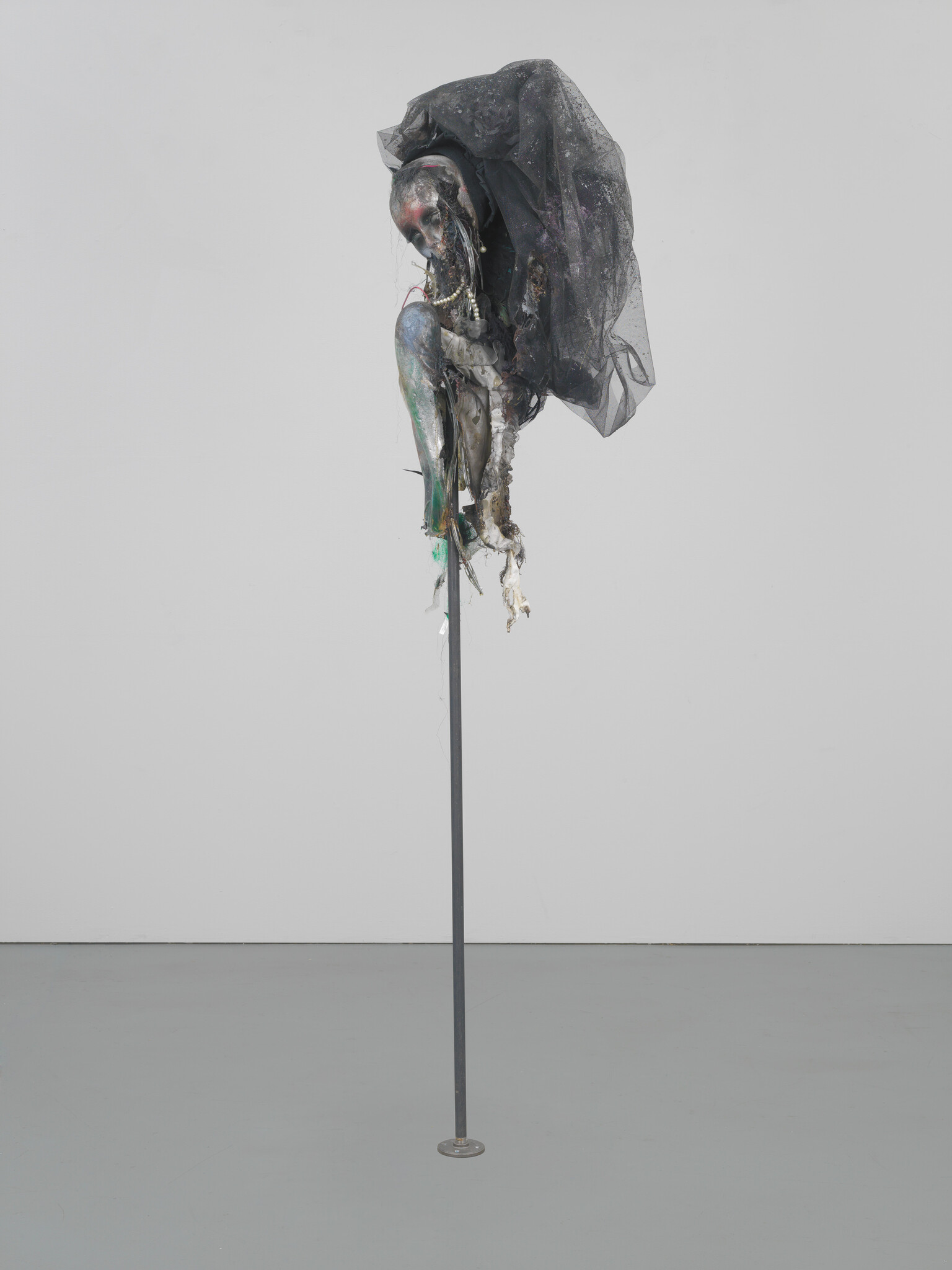 A tattered mannequin head and torso draped in black netting mounted on a tall metal pole.