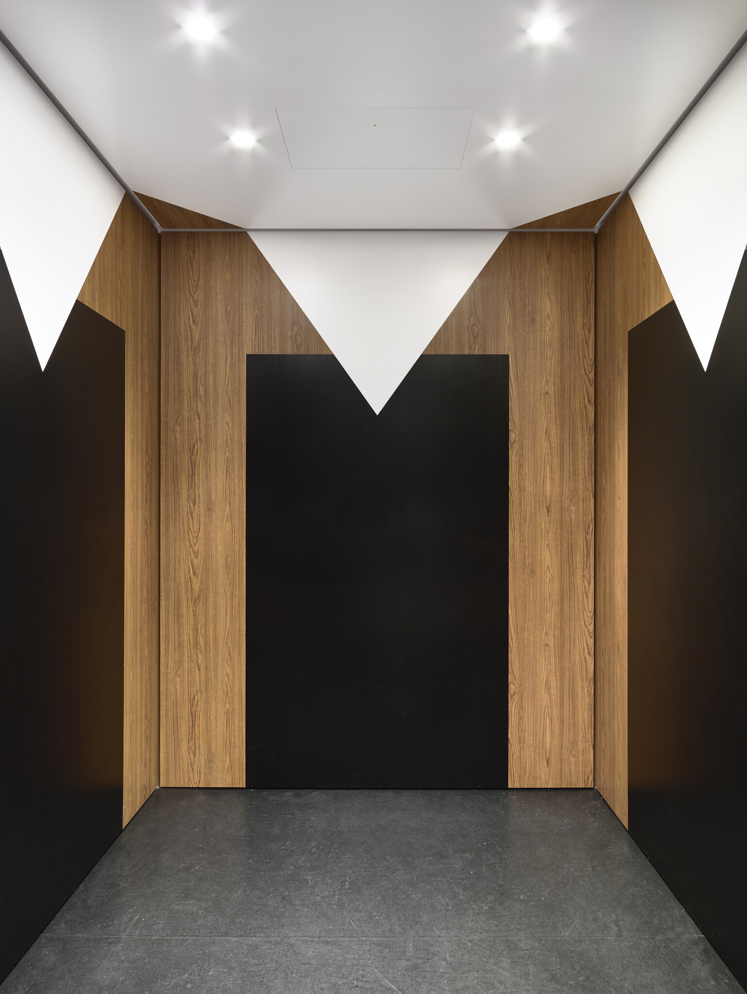 An elevator's interior with geometric wall designs resembling the underside of a table, wood-like columns in the corners for table legs and a white diamond predominating the ceiling for the table cloth