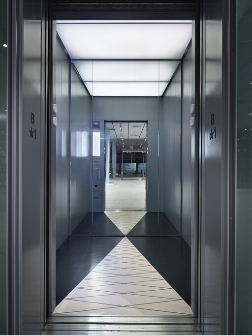 Interior of a modern glass elevator with mirrored walls and a triangular patterned floor.