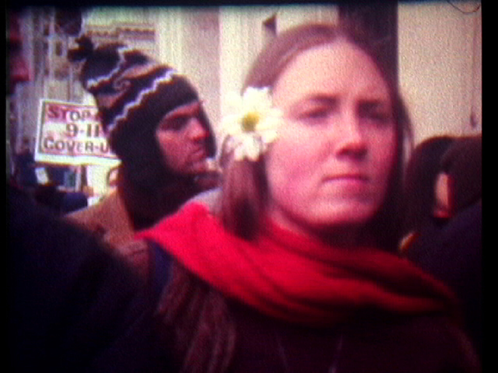 Woman with a daisy in her hair and red scarf stands in a crowd near a "Stop 9-11 cover-up" sign.