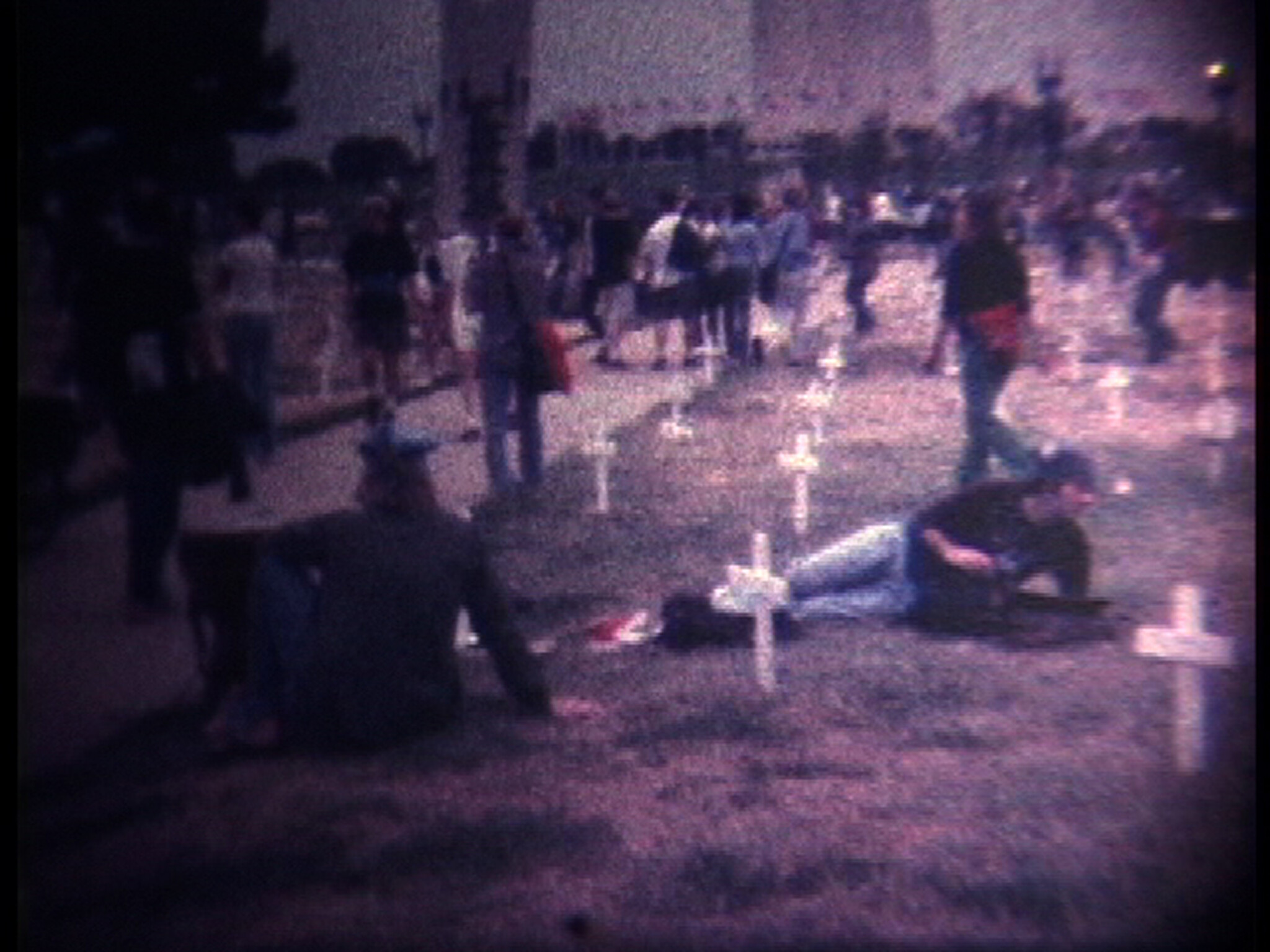 People sit and lie among rows of white crosses in a cemetery while others walk past.