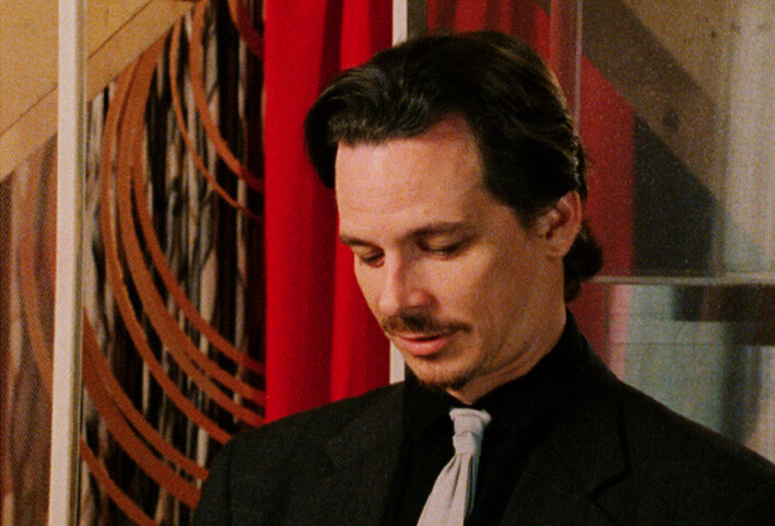 A man in a suit and tie looks down thoughtfully against a red curtain and patterned wall.