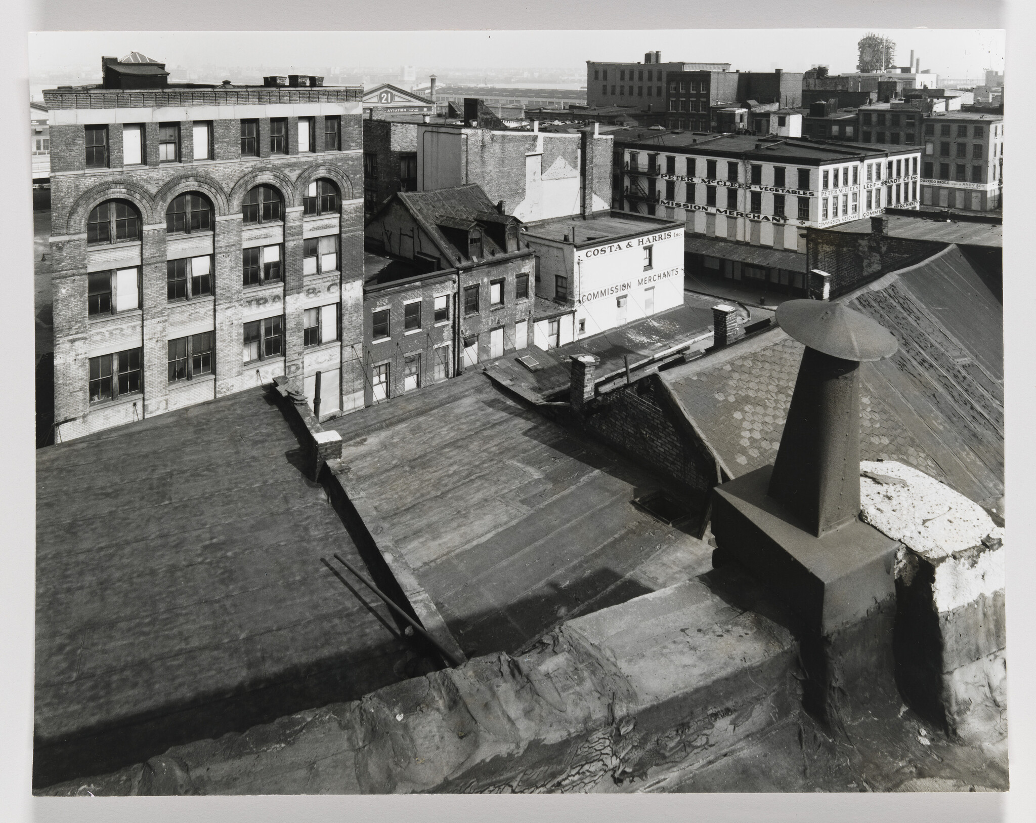 Rooftop chimney and flat roof overlooking brick warehouse buildings with "Costa & Harris" signage.
