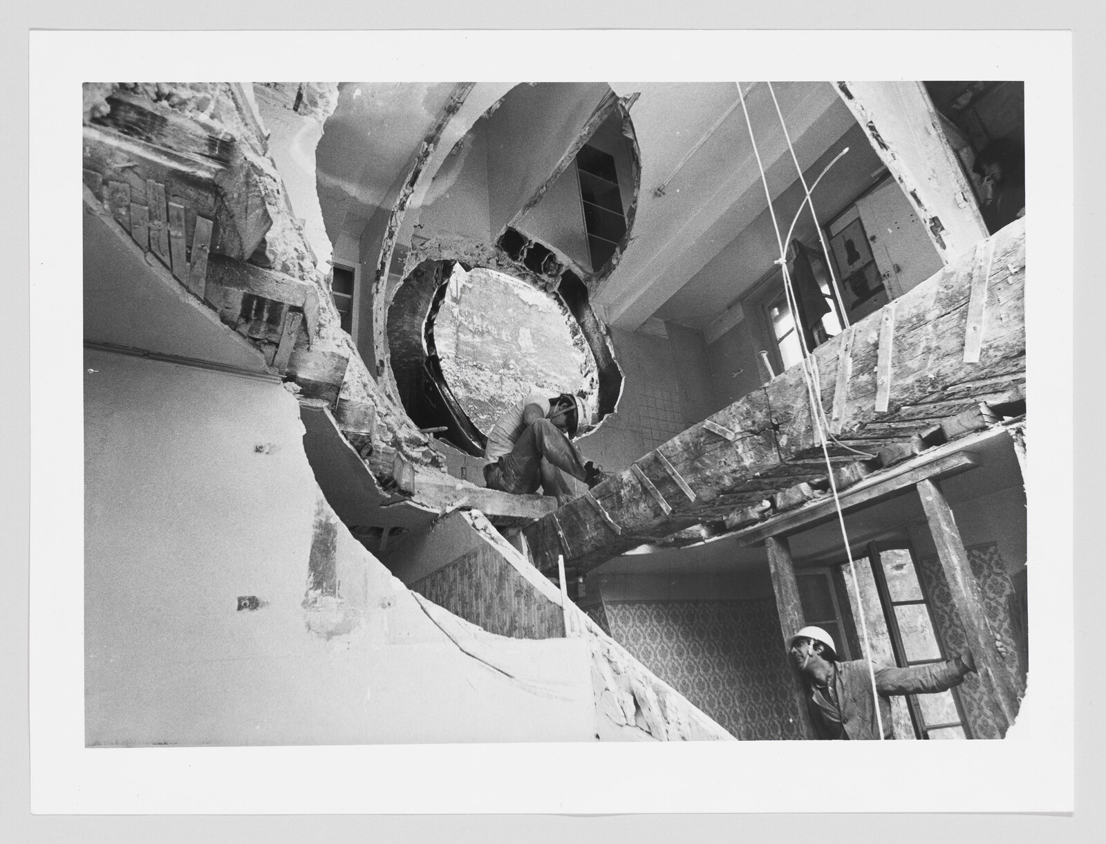 Two construction workers dismantle an interior floor around a large circular opening.