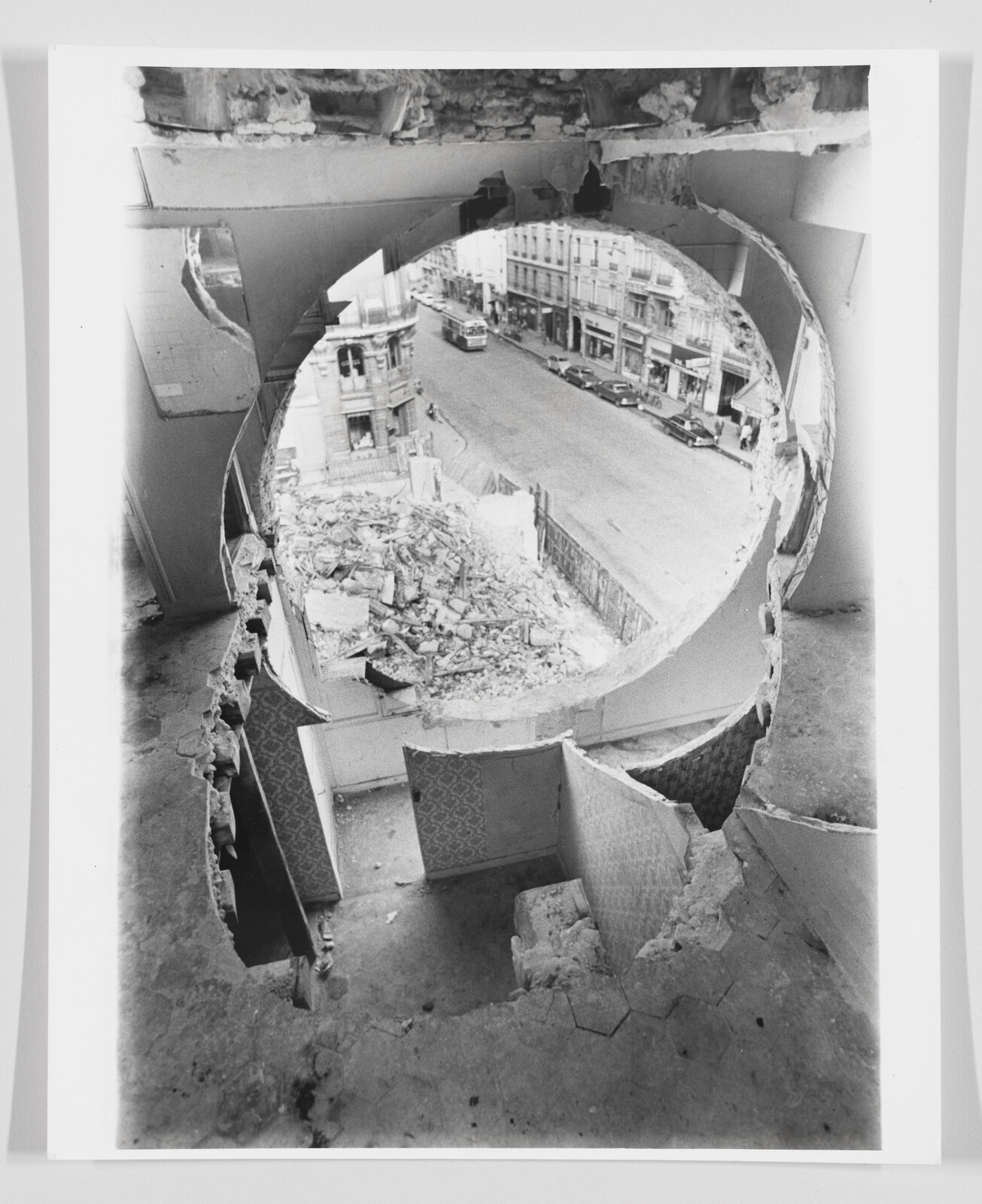 View through a large circular hole in a demolished building showing rubble and a city street below.