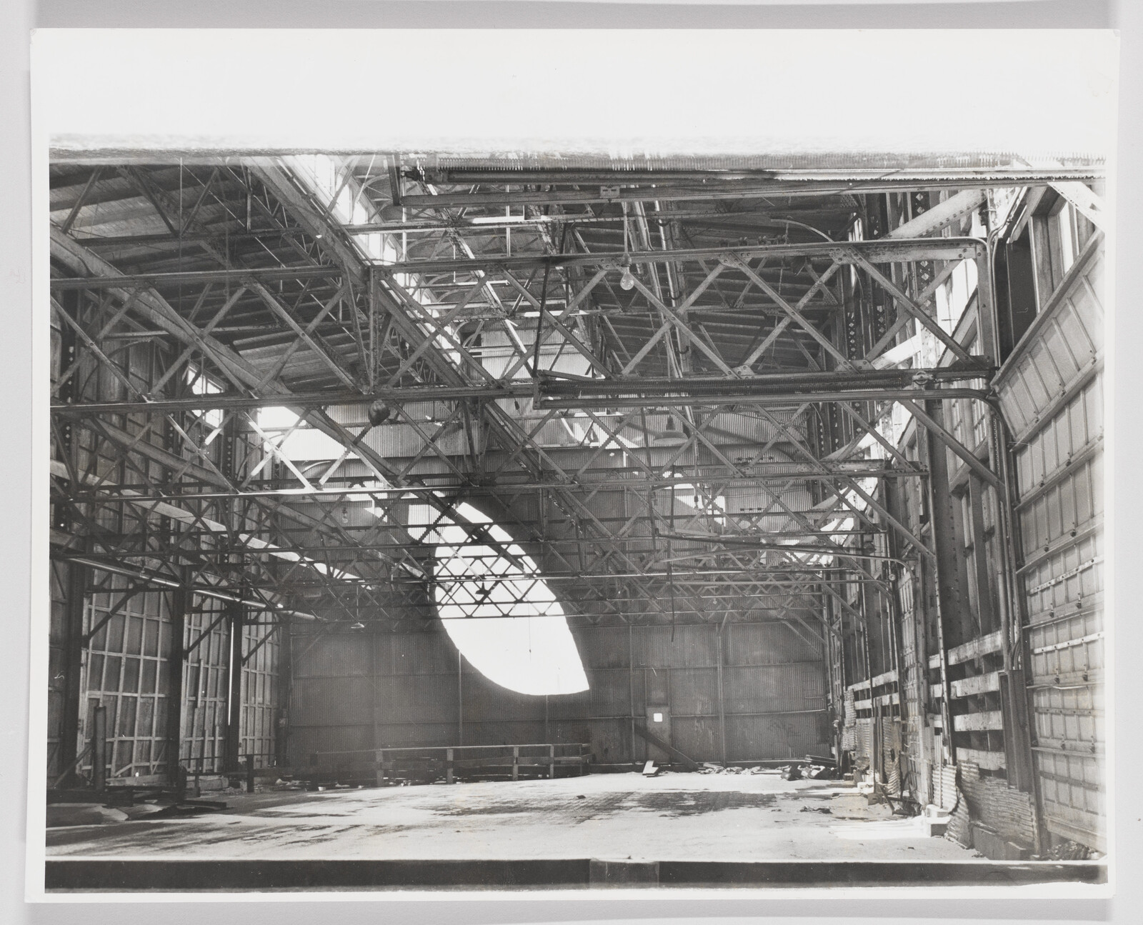 A cavernous, abandoned structure with exposed metal beams and a monumental, leaf-shaped aperture cut into the far wall and filling the space with sunlight.
