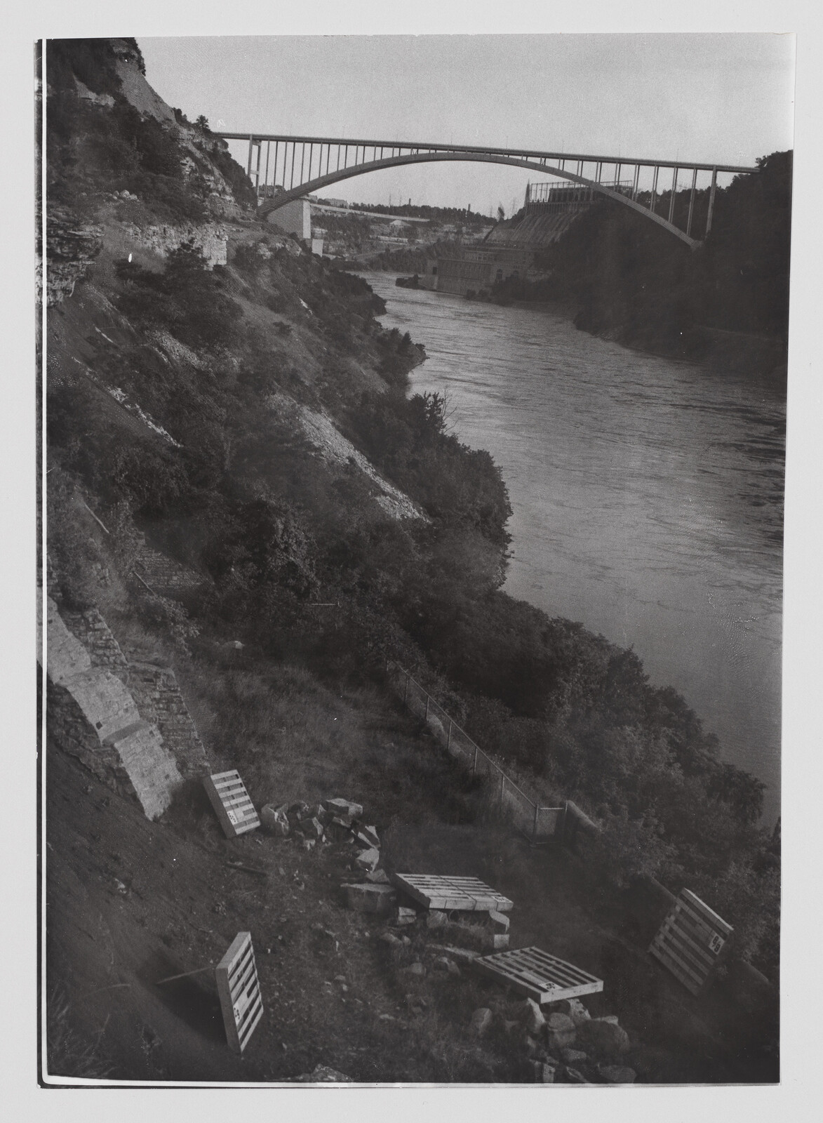 A large arch bridge spans a river through a steep, vegetation-covered canyon with scattered wooden pallets.