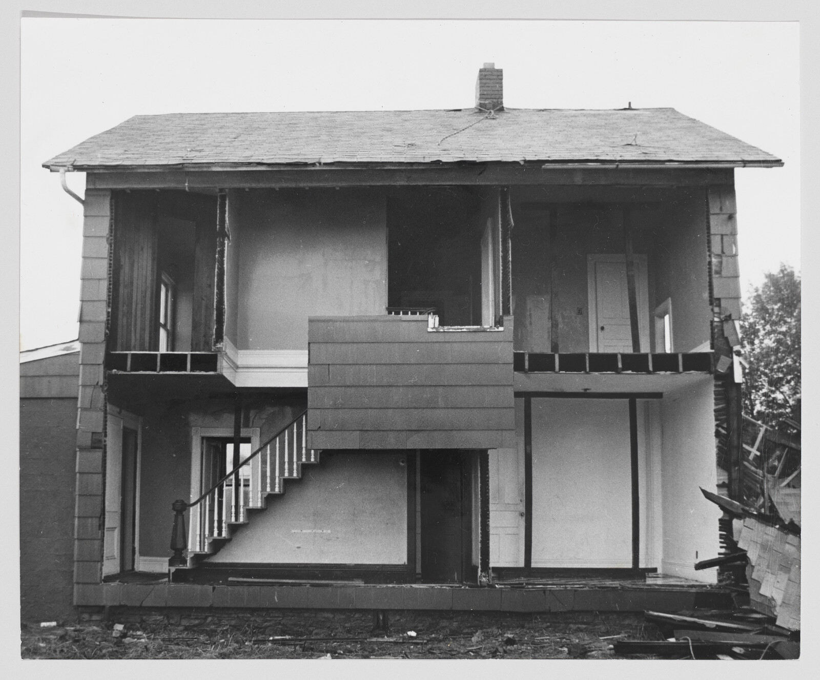 A black and white photograph of a two-story house with its front wall removed, exposing the interior rooms and staircase. The house appears to be in a state of disrepair or under construction, with debris scattered in front of the building.