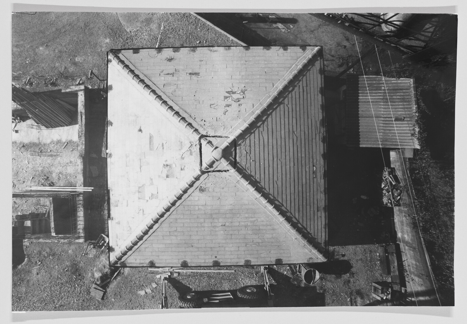 Square building roof with intersecting ridges seen from above, surrounding yard and parked trailer.