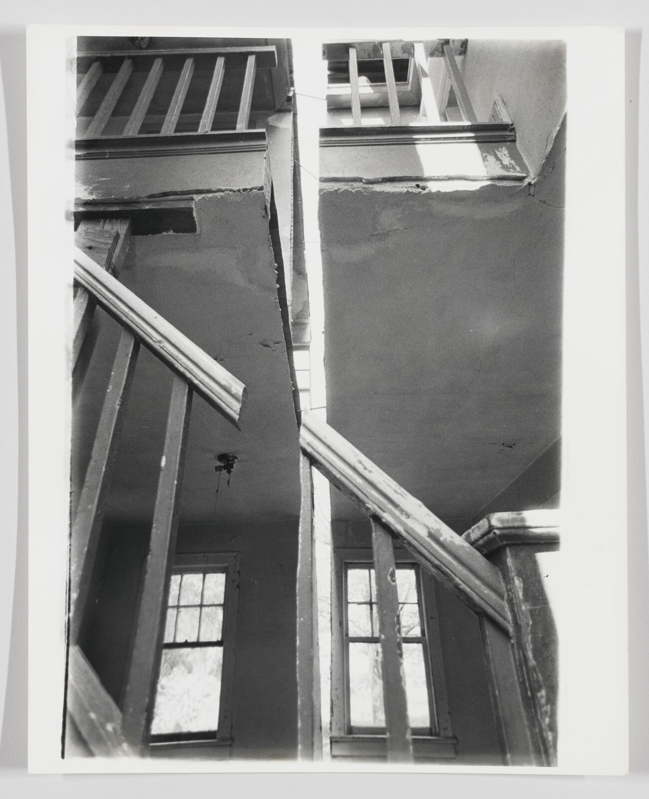 Worn wooden staircase railing leads up through a split ceiling with two tall windows below.