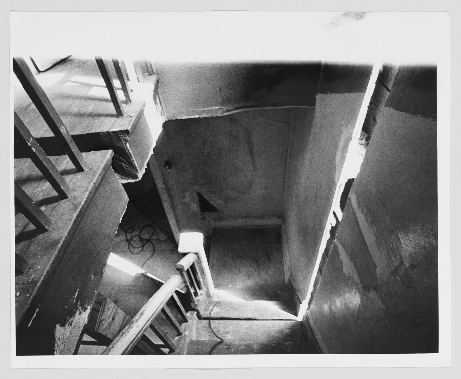 Worn wooden stairwell viewed from above with peeling paint, sunlight along a crack, and a coiled cord.