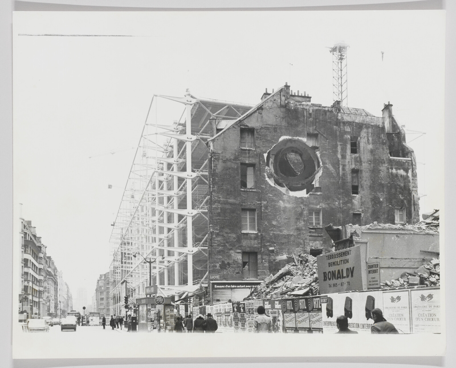 A black and white photograph showing a city street with a partially demolished building on the right, featuring a large circular hole through its remaining facade. Scaffolding structures are visible on the left side of the building. In the foreground, pedestrians walk along the sidewalk, and various signs, including one for a demolition company, are seen amidst the rubble.