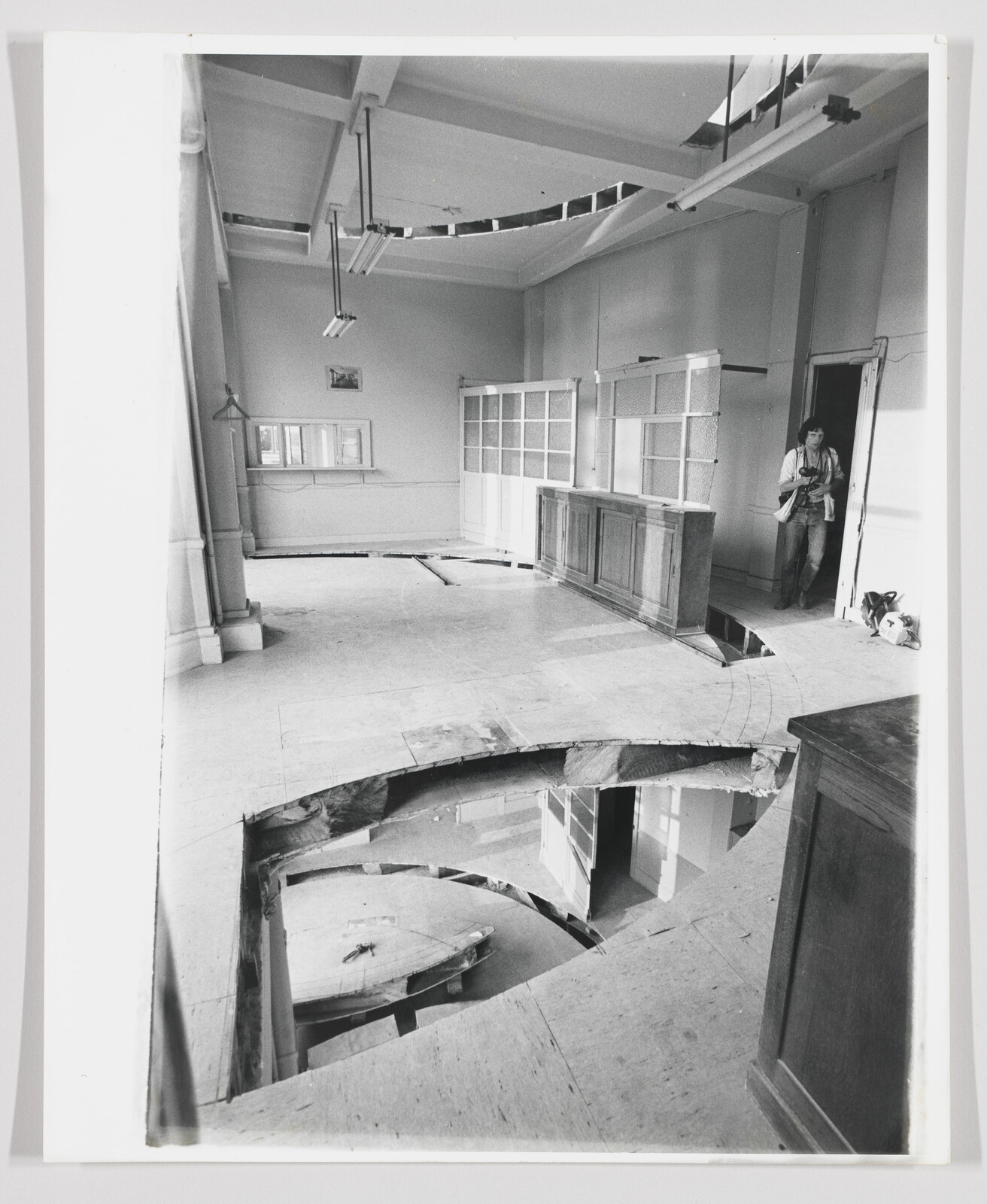 Large circular hole in the wooden floor reveals lower level while a person stands at the doorway.