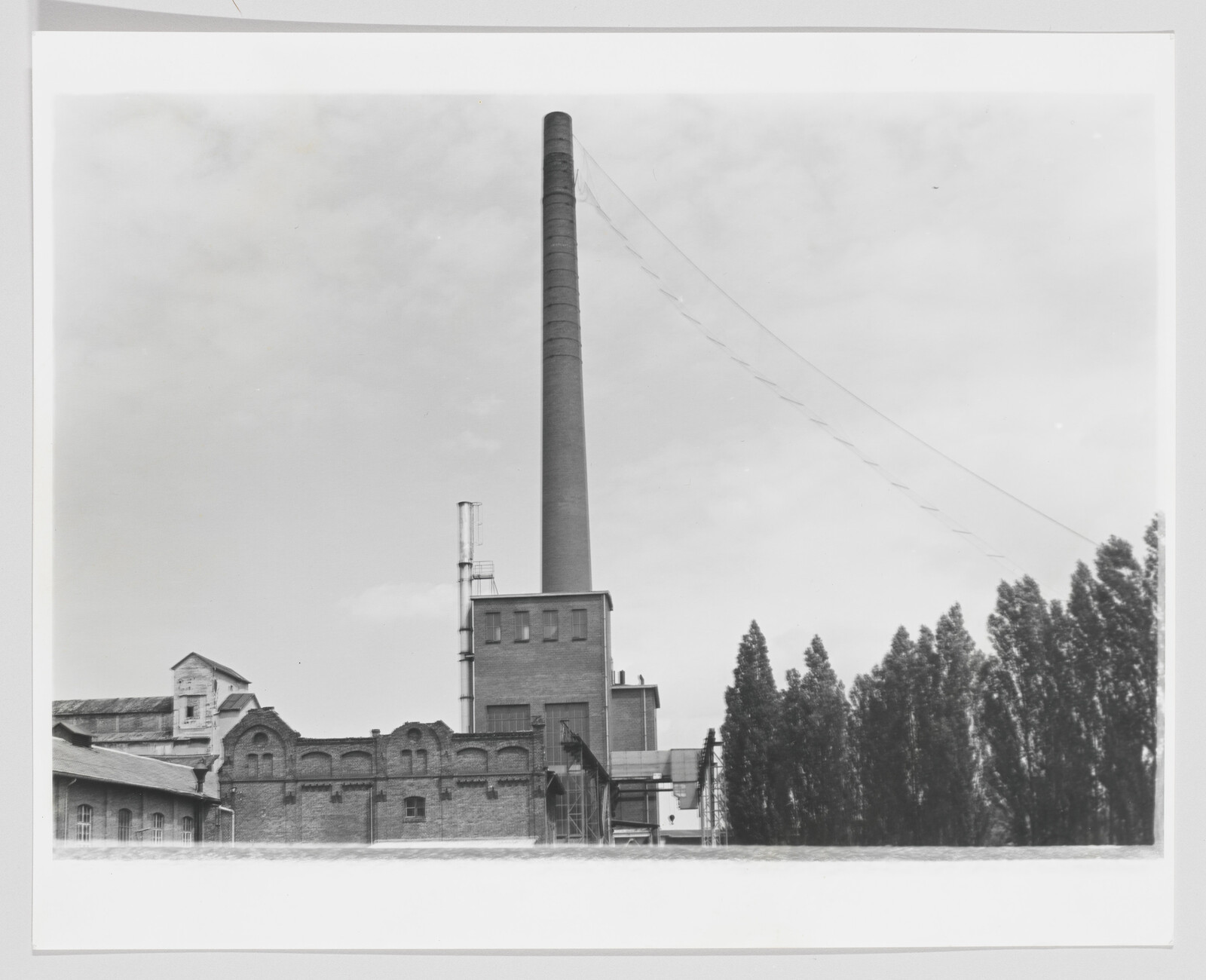 A tall factory smokestack rises behind brick industrial buildings with a row of trees beside them.