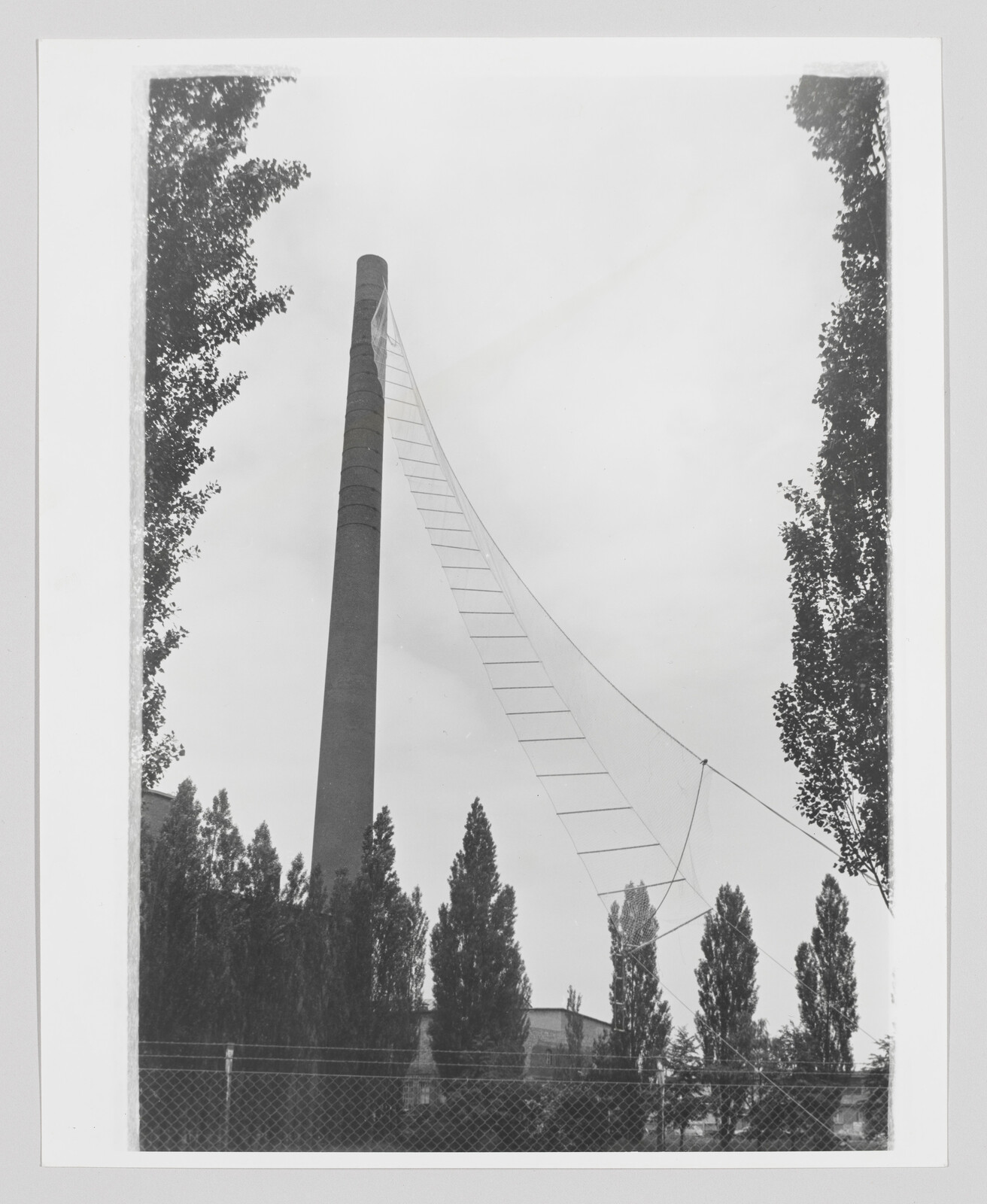 Tall industrial smokestack with a long triangular ladder-like net stretched from its top toward the ground.