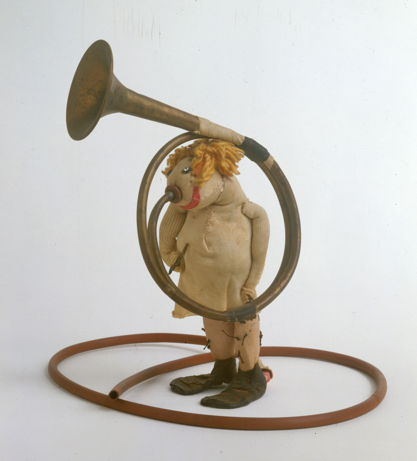 A worn cloth doll stands with a brass horn coiled around its body and feet.