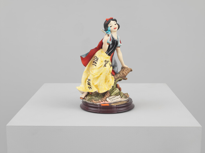 Snow White figurine wearing a yellow skirt and red cape sits on a wooden base holding a blue bird.