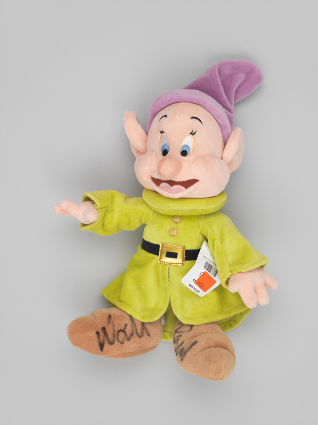 A plush dwarf wearing a green coat and purple hat with signatures on its feet and a price tag.