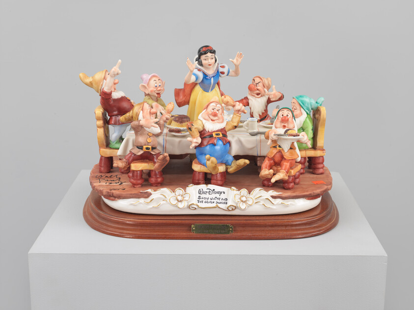 Snow White stands behind a table as the seven dwarfs eat, cheer, and pose around her.
