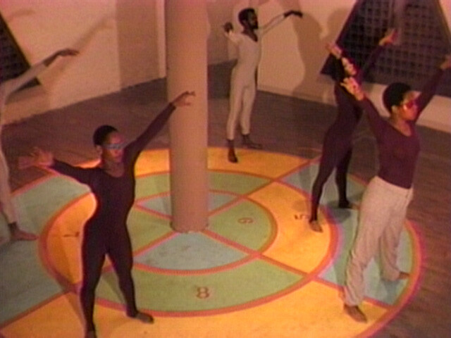 Five people in leotards stand on a colorful numbered floor, stretching their arms upward around a central pillar.
