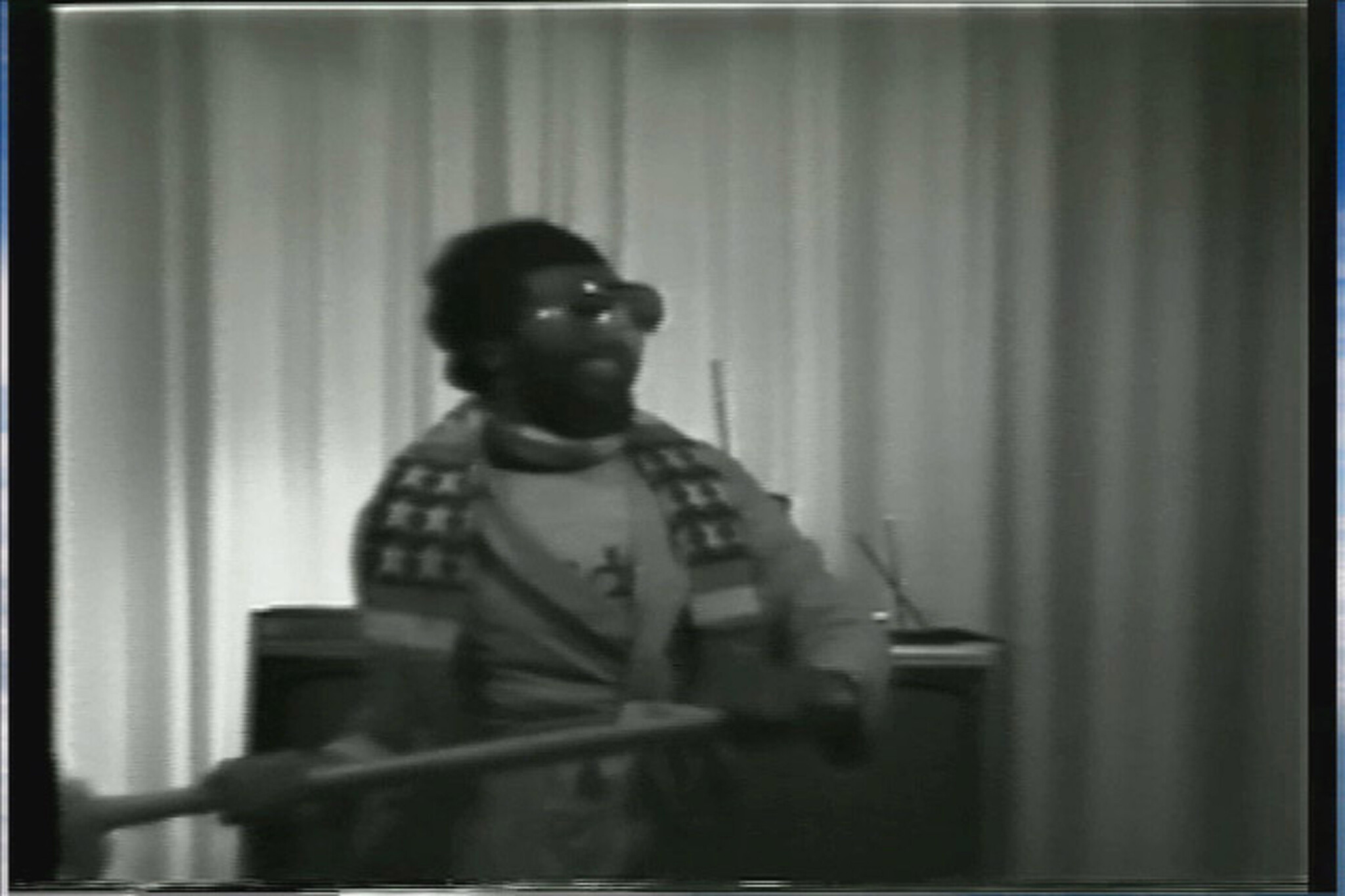 Man wearing glasses and a patterned scarf holds a long pole while standing in front of curtains.