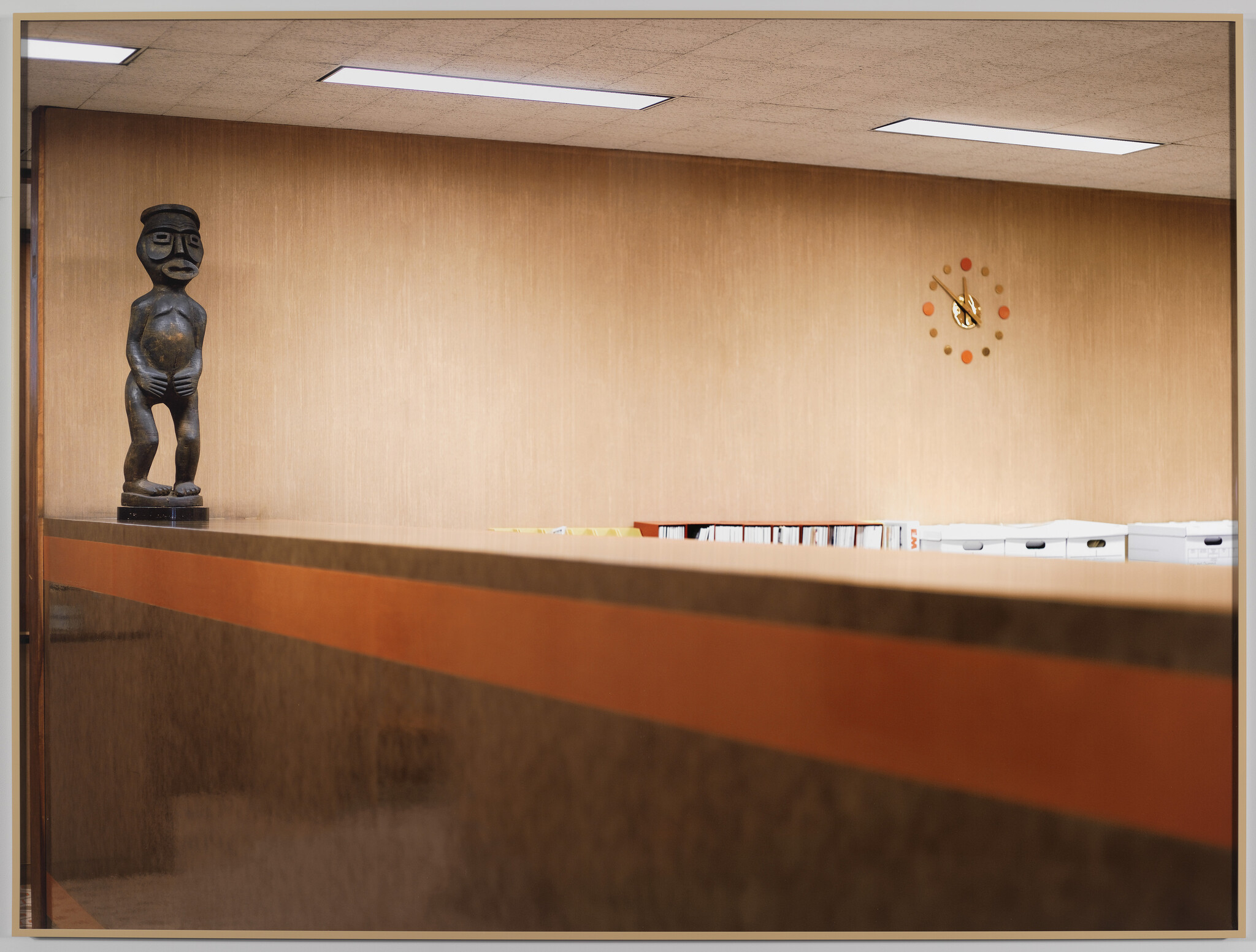 A wooden tribal statue stands on a reception counter in a quiet office with a wall clock.