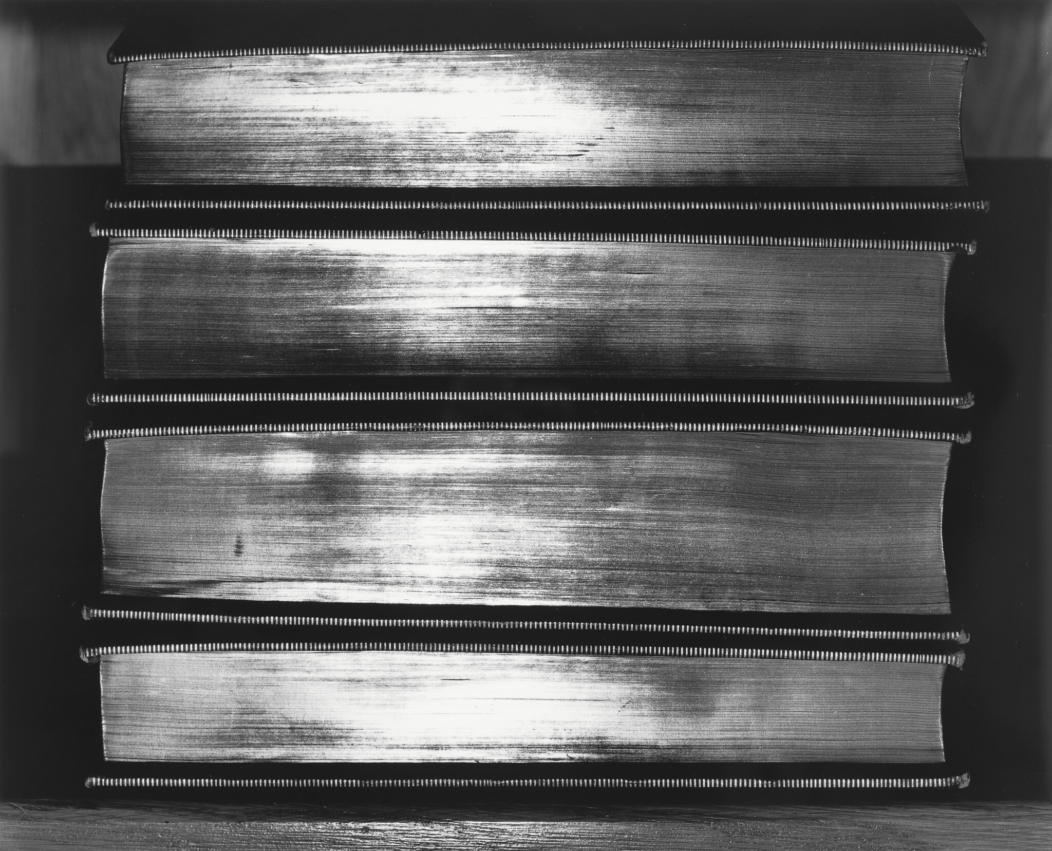 Four large thick books stacked horizontally with shiny, textured fore-edges visible.