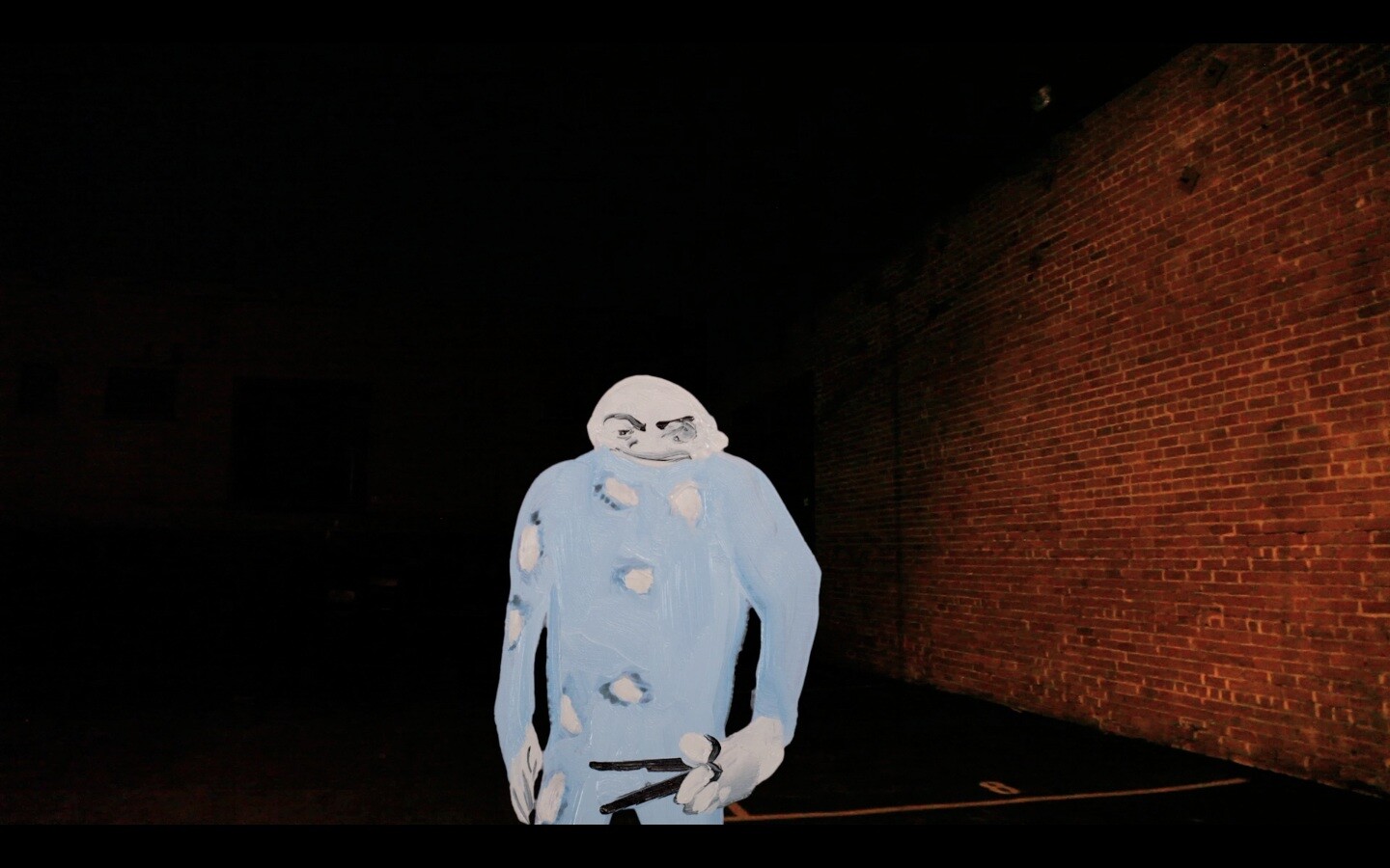 A person in a costume that resembles a cartoonish ghost with a grumpy expression stands in a dark alley with a brick wall in the background. The ghost costume is blue with multiple holes, and the person's face is visible through the head opening, painted to match the costume.