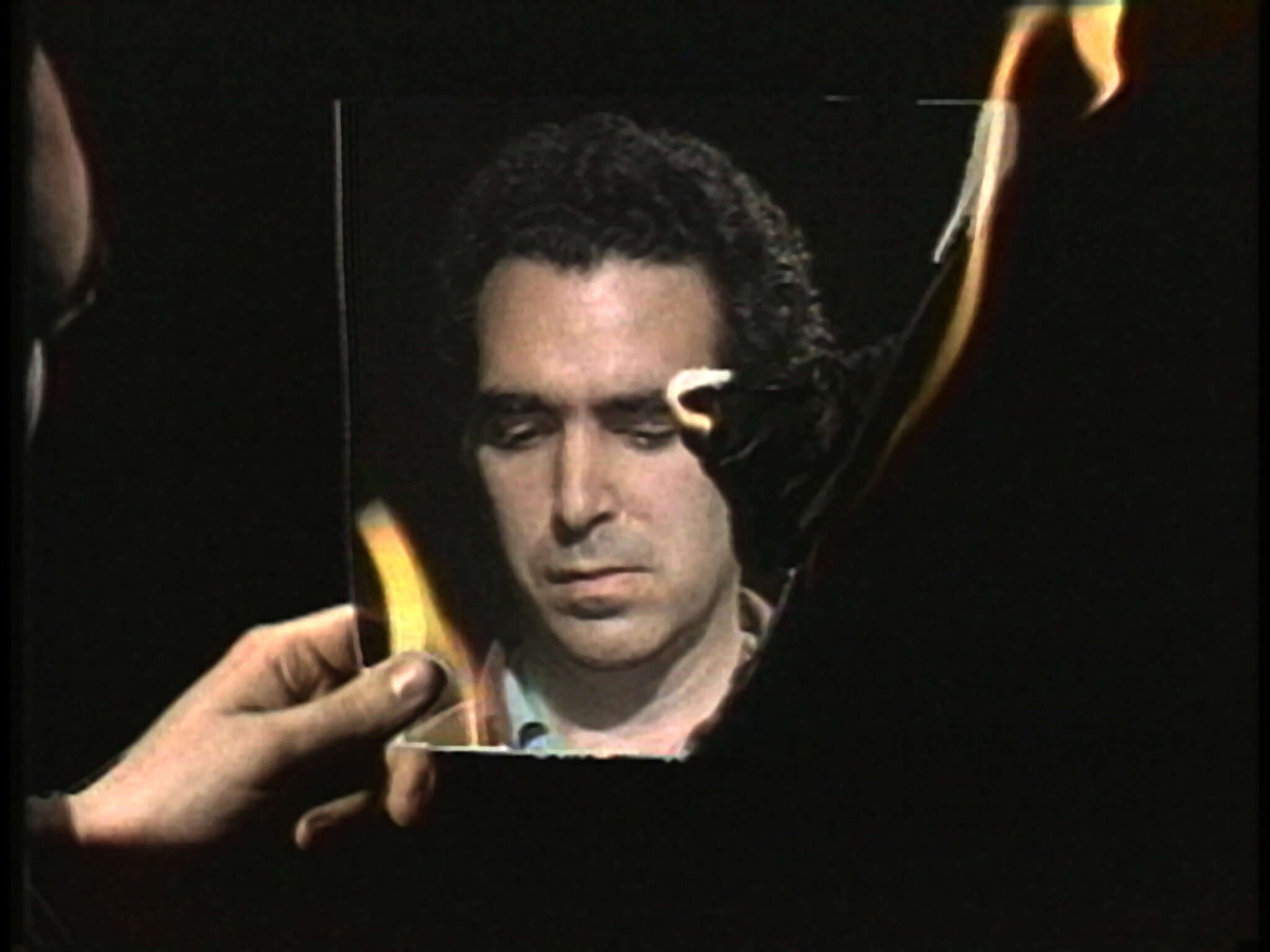 A hand holds a burning photograph showing a man's solemn face as flames consume the paper.