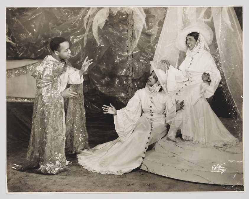 A vintage black and white photograph depicting three individuals in theatrical costumes on a stage set. On the left, a person wearing a shiny, silver cape and trousers gestures towards the center, where another individual is kneeling on the ground, dressed in a flowing white gown and a headpiece, looking up with an expressive pose. To the right, a third person stands with one hand raised, also clad in an elaborate white costume with a wide-brimmed hat, engaging with the central figure. The backdrop features metallic-looking foliage, and the overall scene suggests a dramatic moment in a play or performance.