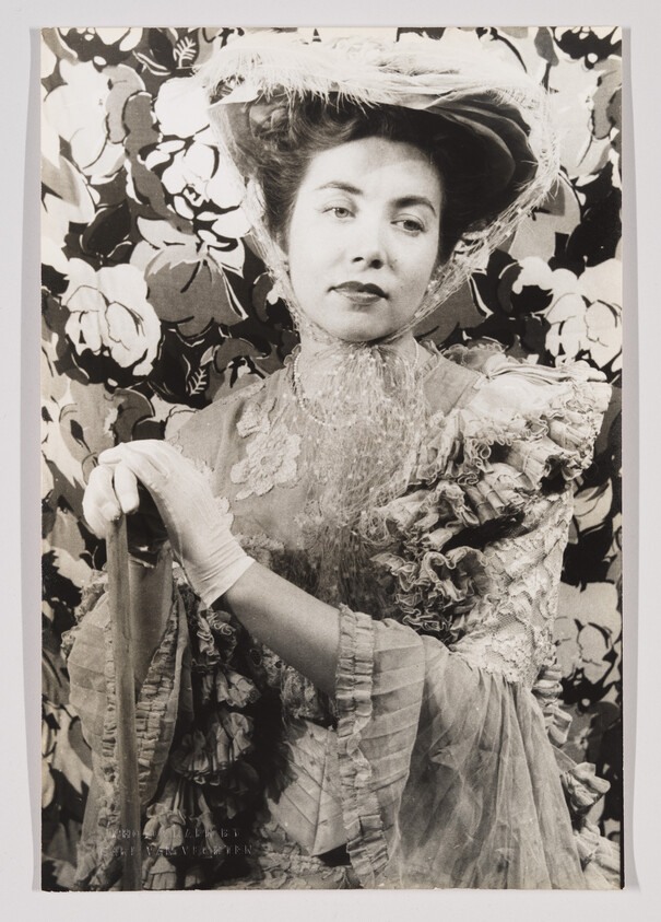 A black and white photograph of a woman dressed in vintage clothing, featuring a wide-brimmed hat and a lace-trimmed dress, posing in front of a floral backdrop.