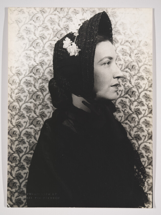 A vintage black and white photograph of a woman in profile. She is wearing a dark, textured headscarf adorned with a white flower and a dark coat. The background features a floral patterned wallpaper. The bottom of the image includes text that attributes the photograph to Carl Van Vechten.