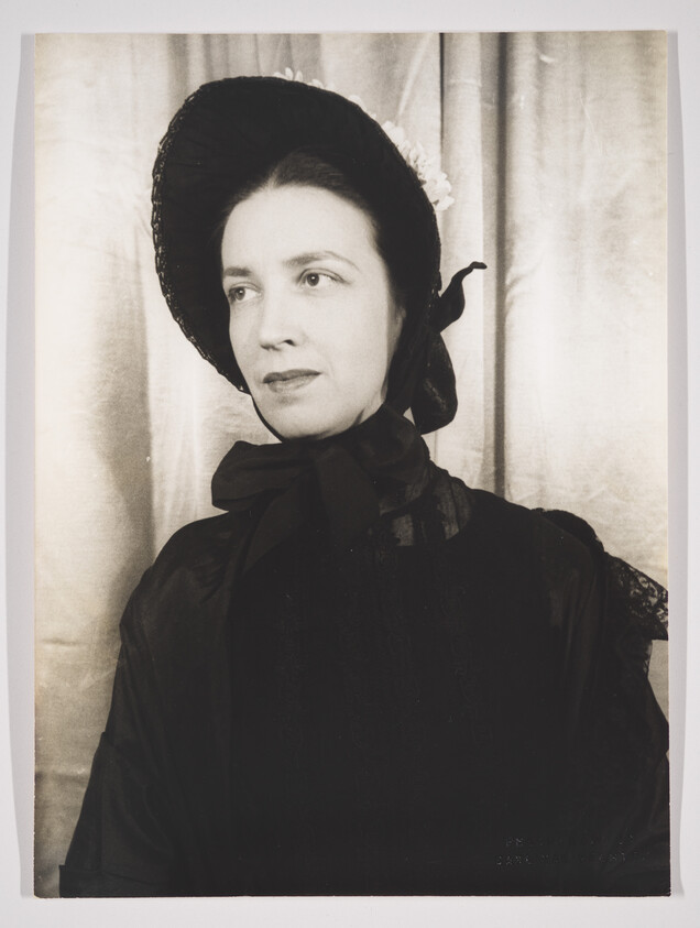 Vintage black and white portrait of a woman wearing a wide-brimmed hat adorned with flowers and a dark, high-collared dress with a bow at the neck. She has a contemplative expression and is looking off to the side, with a neutral backdrop that resembles draped fabric.