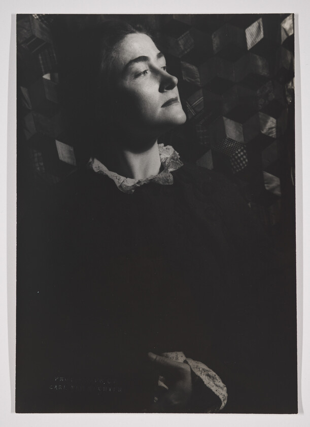 A black and white photograph of a woman in profile with her eyes closed, featuring dramatic lighting that casts shadows on her face. She wears a dark dress with a lace collar and is set against a background of geometric patterns.