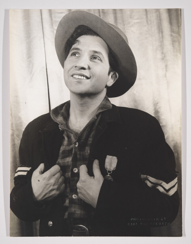 A black and white portrait of a smiling man looking upwards, wearing a wide-brimmed hat and a dark jacket over a plaid shirt. He has a medal pinned to his jacket and is holding onto the lapels. The background appears to be a draped fabric, and there is a signature at the bottom that reads "Photograph by Carl Van Vechten."
